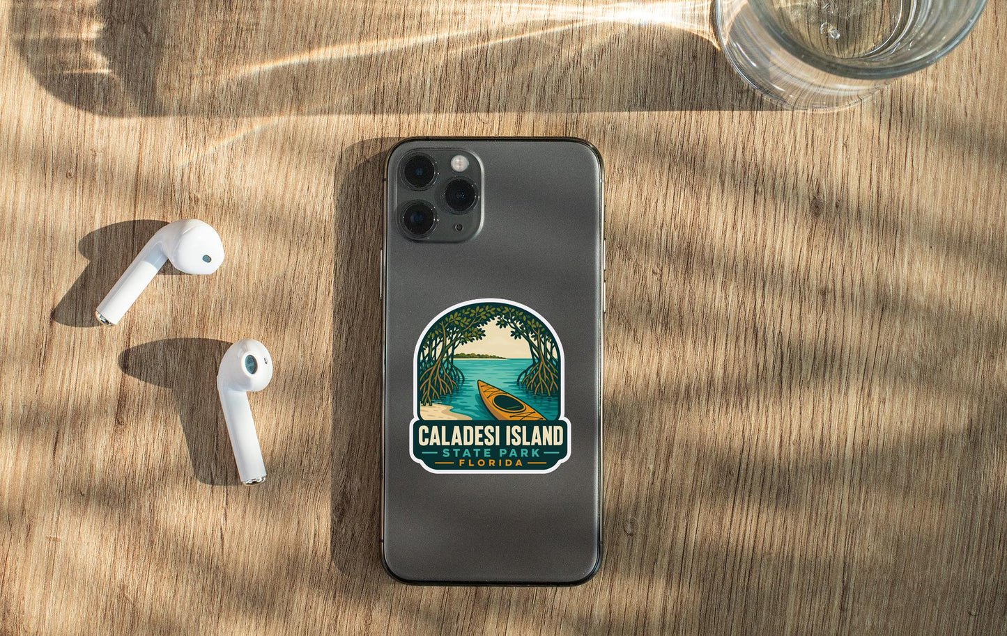 a smartphone with a sticker on the back, placed on a wooden surface alongside a pair of white earbuds and a glass of water.