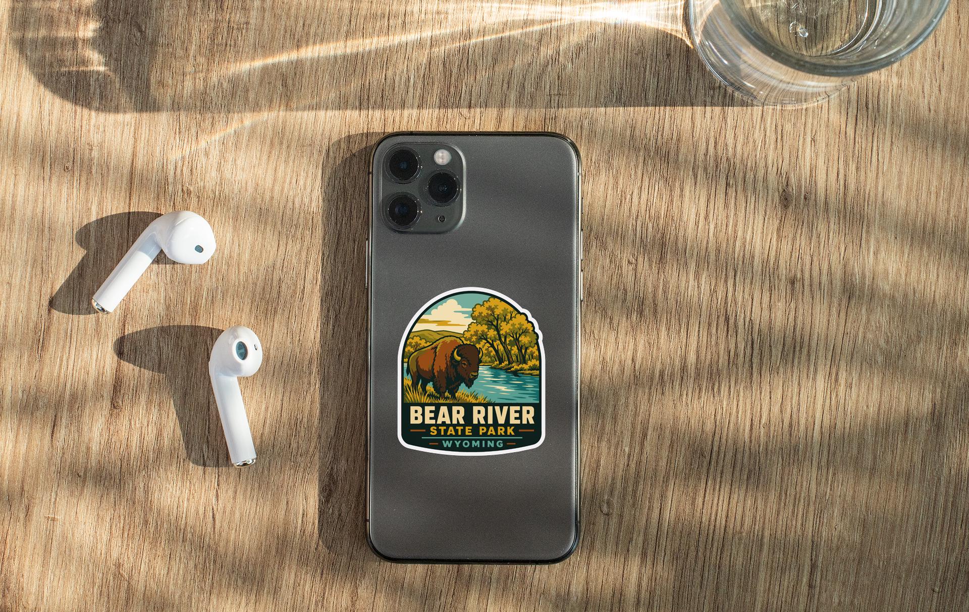 a smartphone with a sticker of Bear River State Park on its back, along with a pair of white earbuds and a glass of water on a wooden surface.