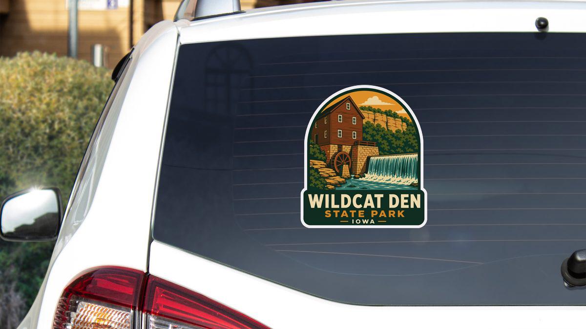 A sticker on the rear window of a white car, depicting a scenic landscape with a waterfall and a building, representing Wildcat Den State Park in Iowa.