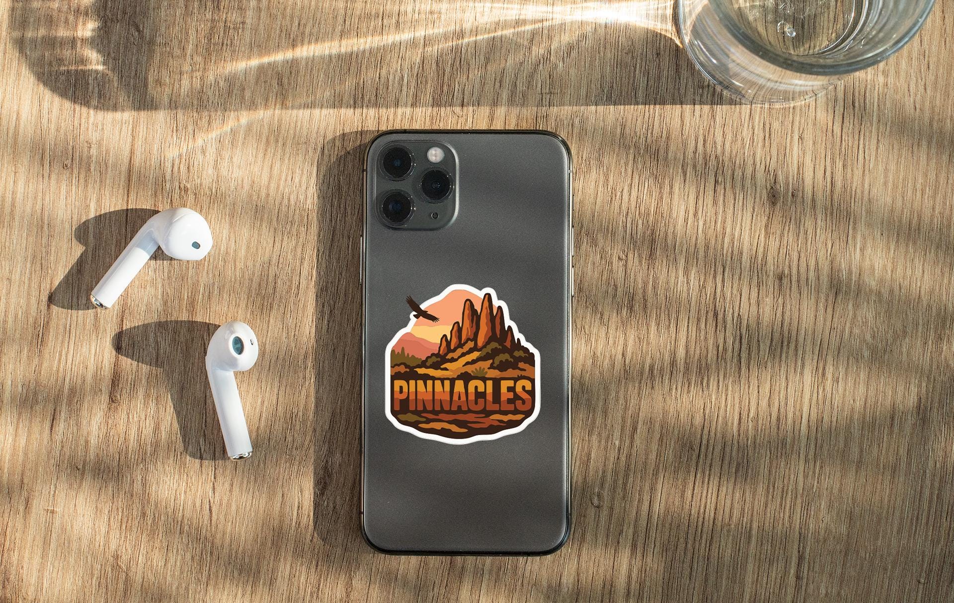a smartphone with a sticker on the back, along with a pair of white earbuds and a glass of water on a wooden surface.