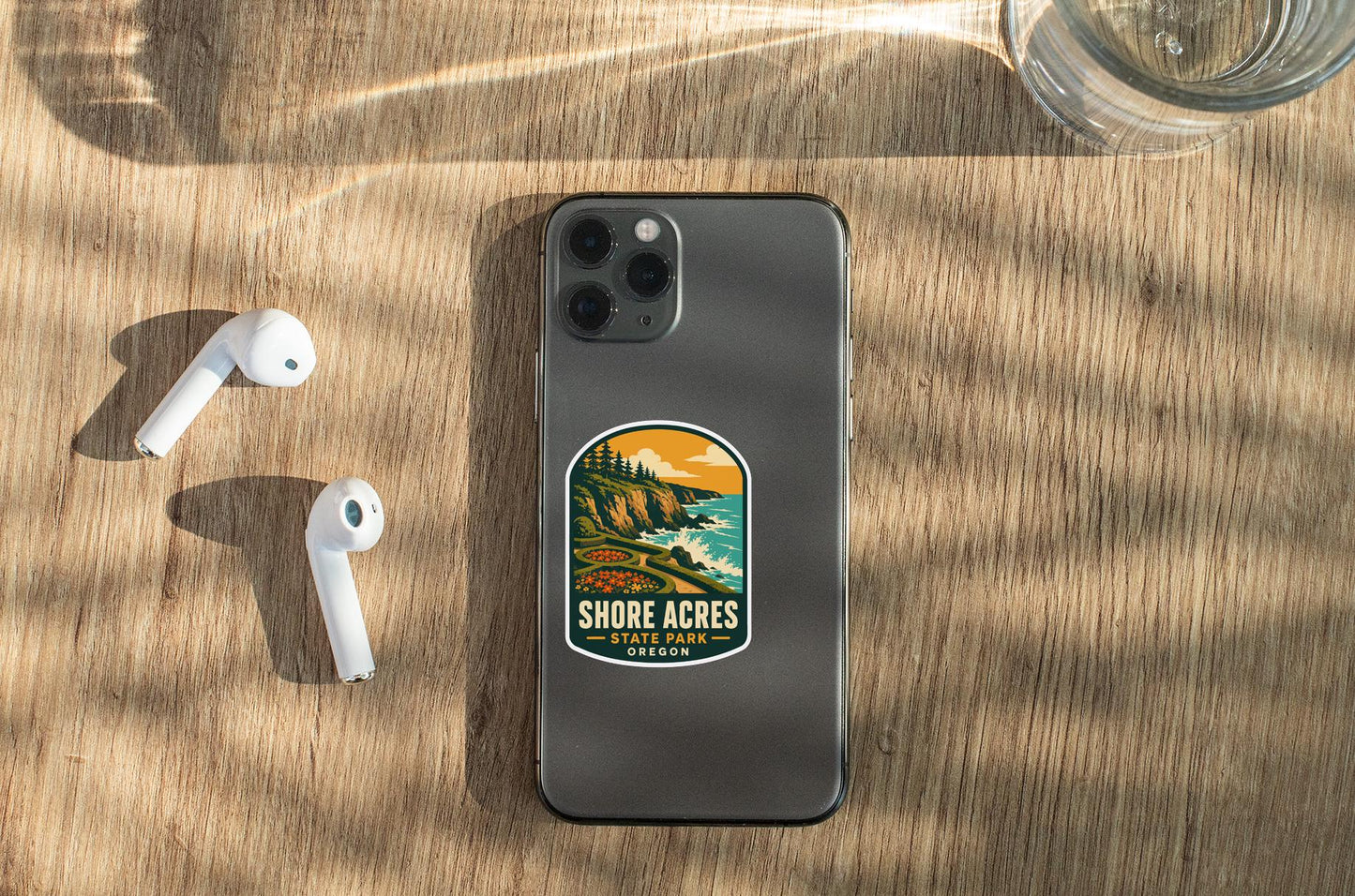 a smartphone with a sticker on the back, placed on a wooden surface alongside a pair of white earbuds and a glass of water.