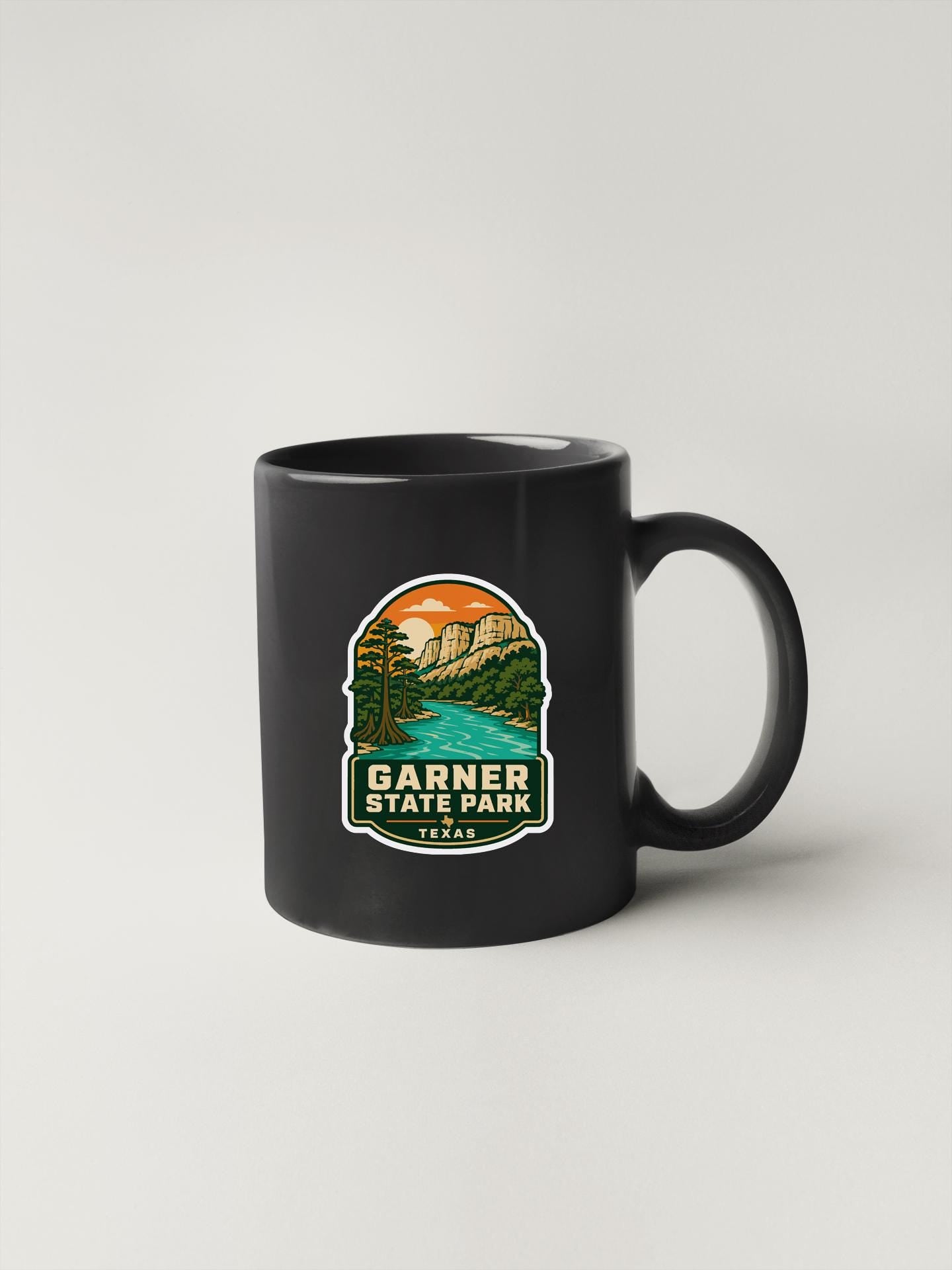 A black coffee mug with a graphic design depicting a scenic landscape, including mountains, a river, and a sunset.