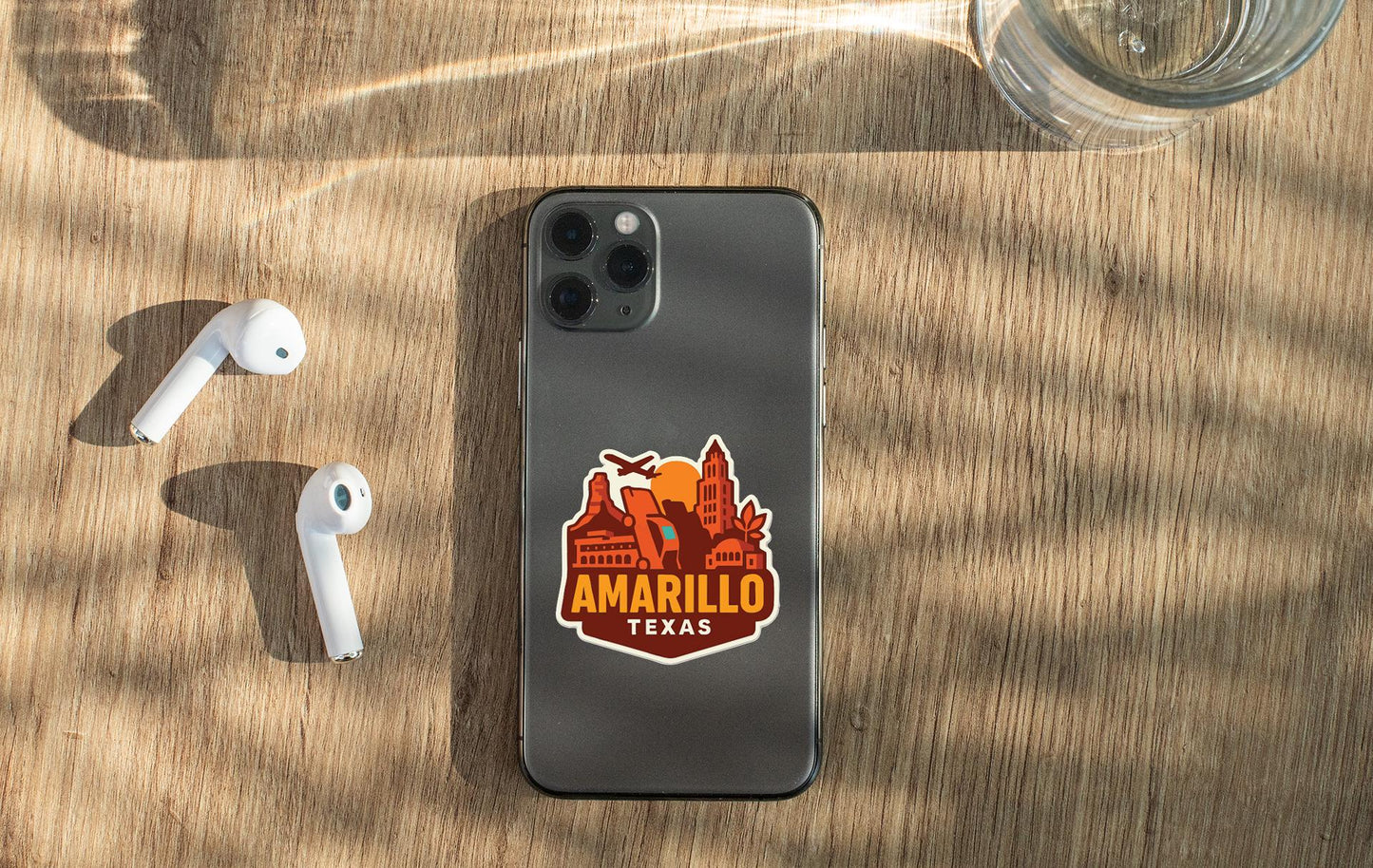 a smartphone with a sticker on the back, placed on a wooden surface alongside a pair of white earbuds and a glass of water.