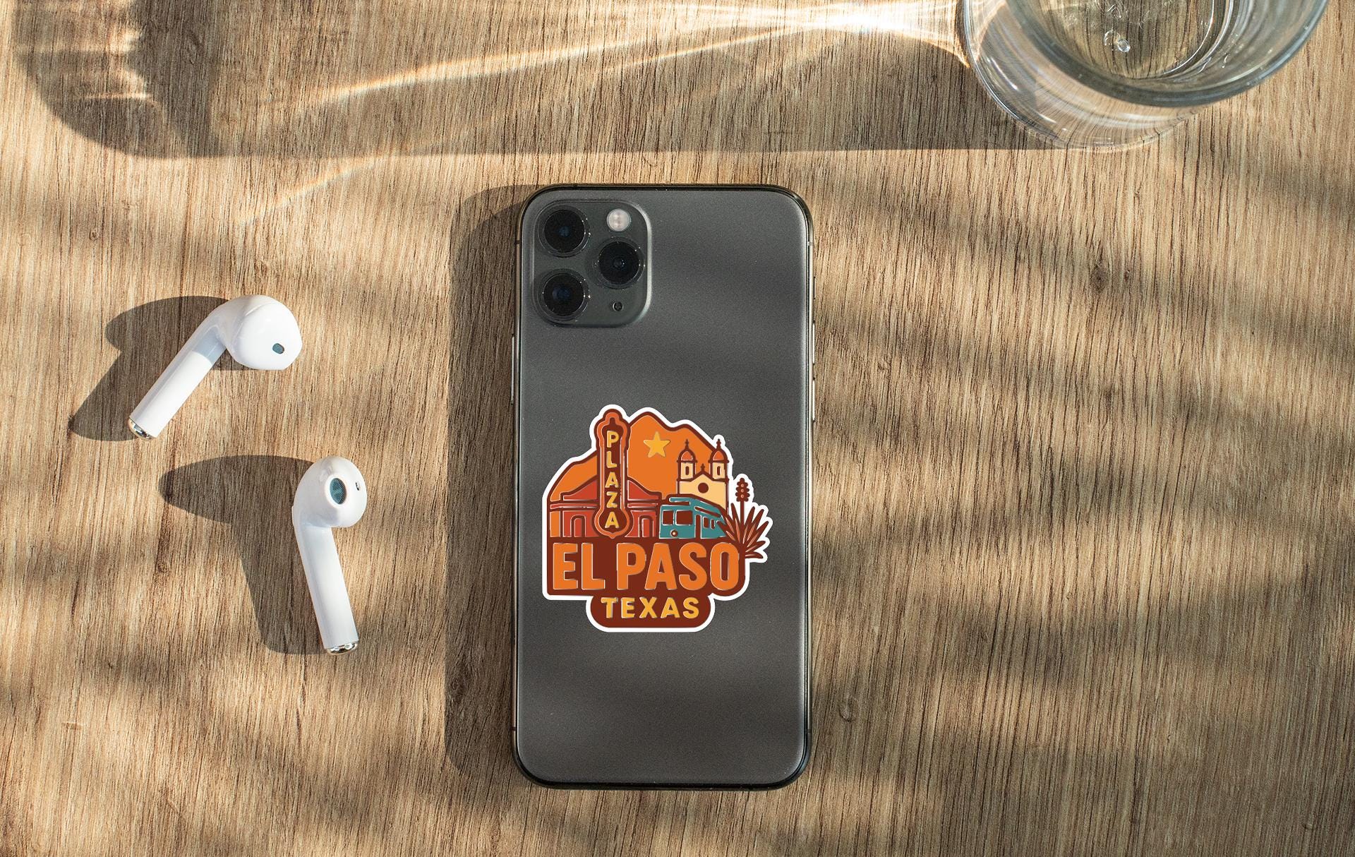 a smartphone with a sticker of El Paso, Texas, placed on a wooden surface alongside a pair of white earbuds and a glass of water.