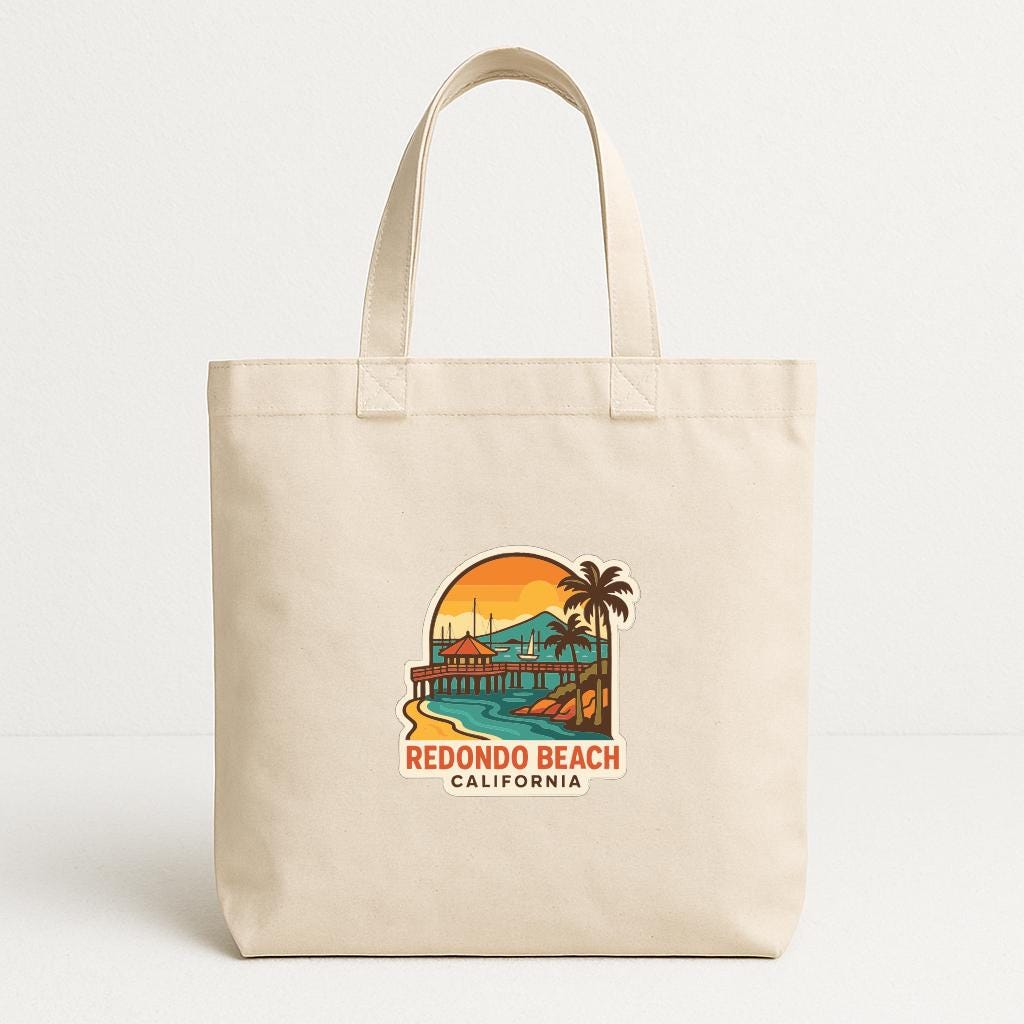 A beige tote bag with a graphic design depicting a beach scene and the text "REDONDO BEACH CALIFORNIA".