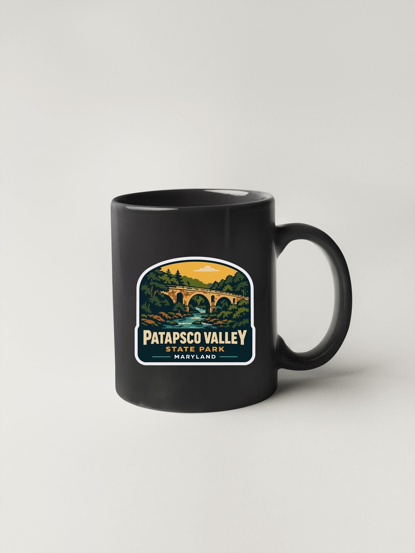 A black coffee mug with a sticker depicting a scenic landscape featuring a bridge and trees.
