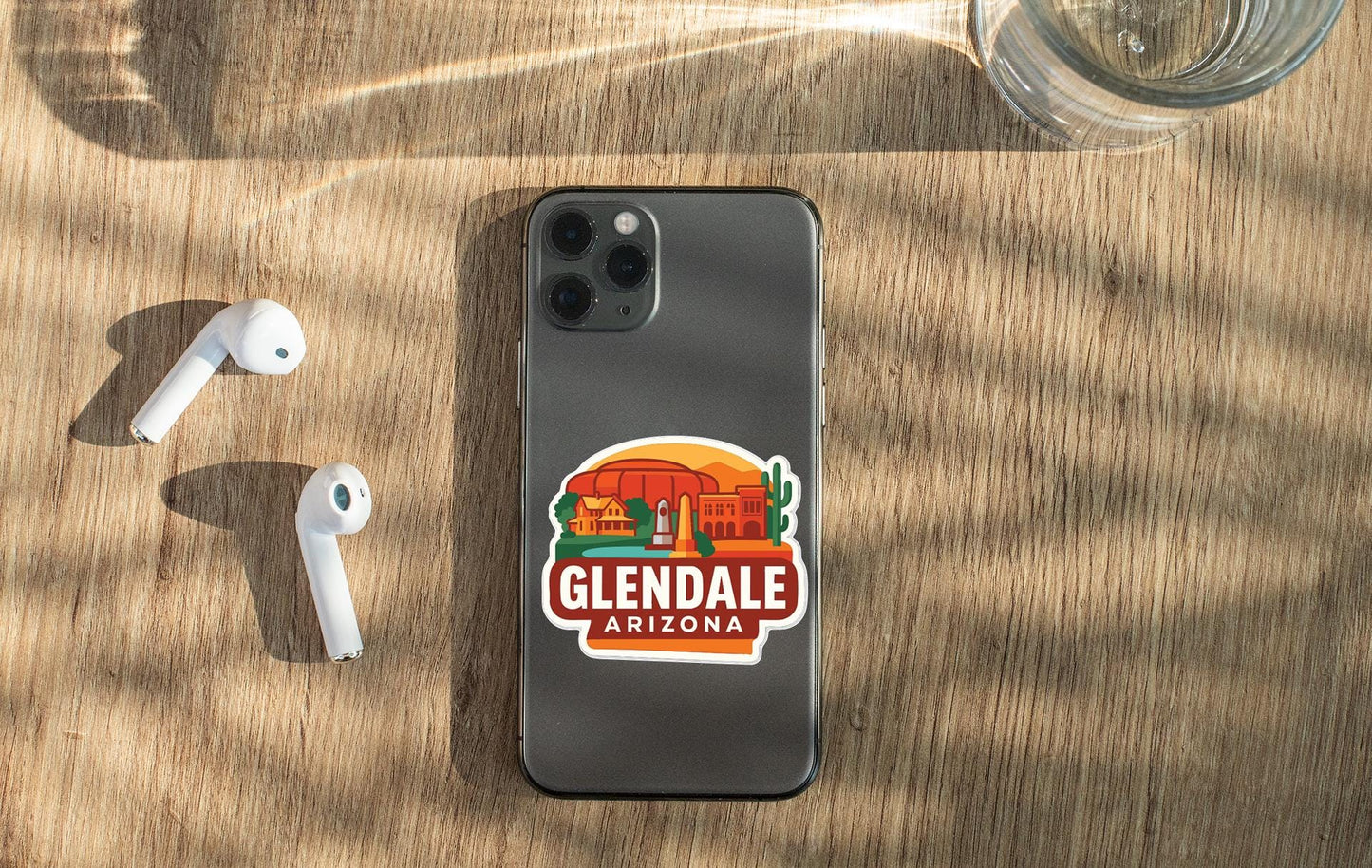 a smartphone with a sticker of Glendale, Arizona, placed on a wooden surface alongside a pair of white earbuds and a glass of water.