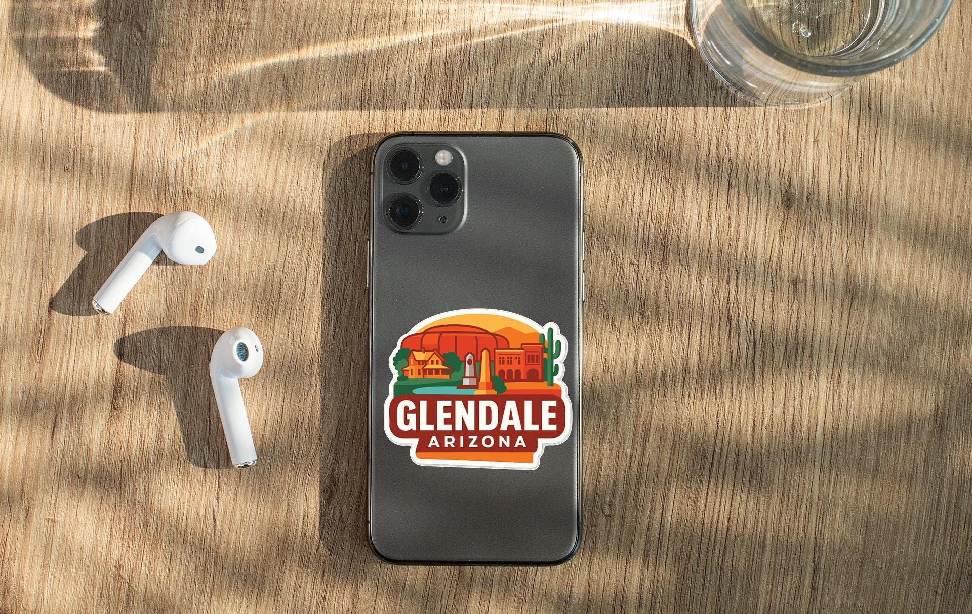 a smartphone with a sticker of Glendale, Arizona, placed on a wooden surface alongside a pair of white earbuds and a glass of water.