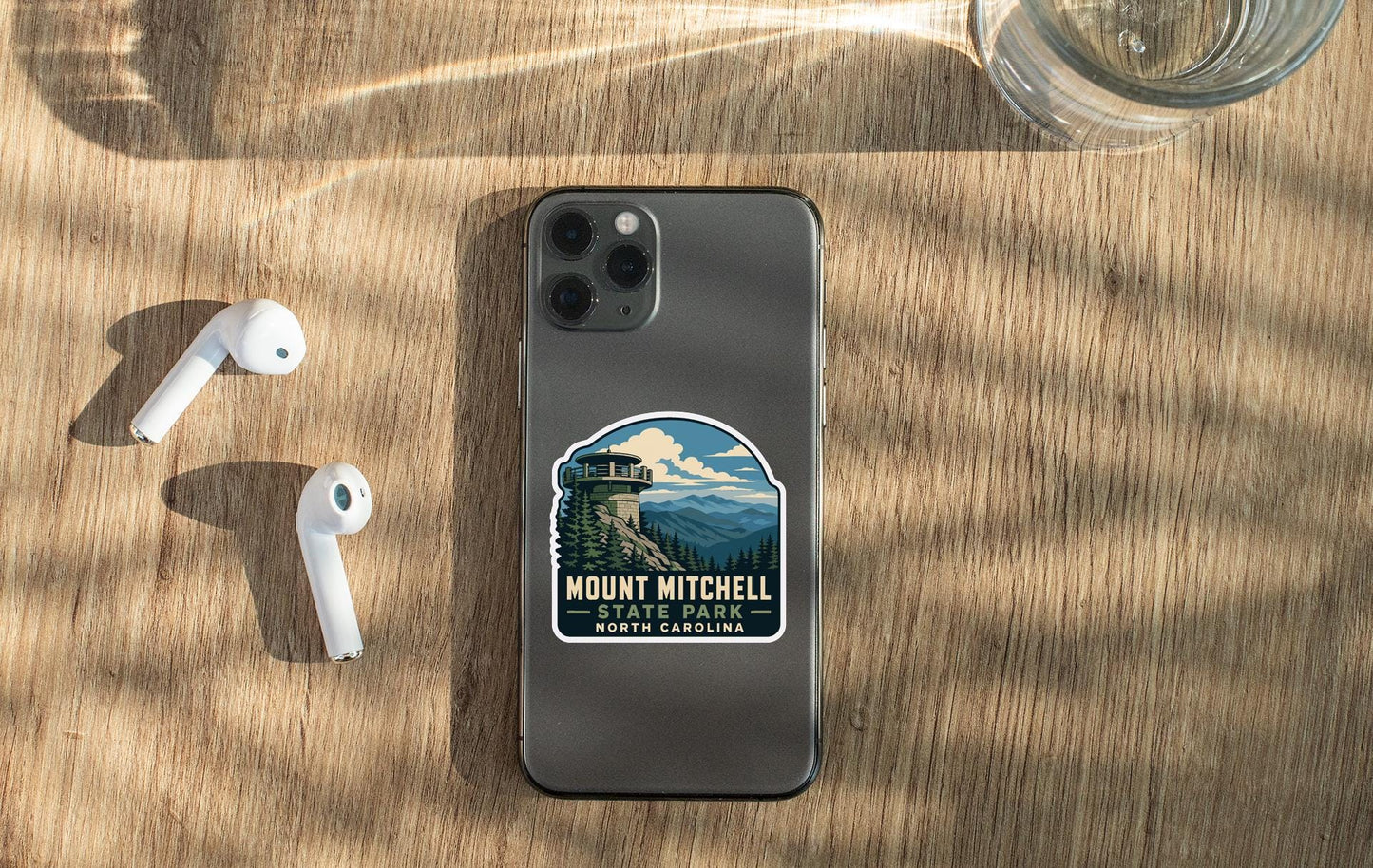a smartphone with a sticker on the back, placed on a wooden surface alongside a pair of white earbuds and a glass of water.