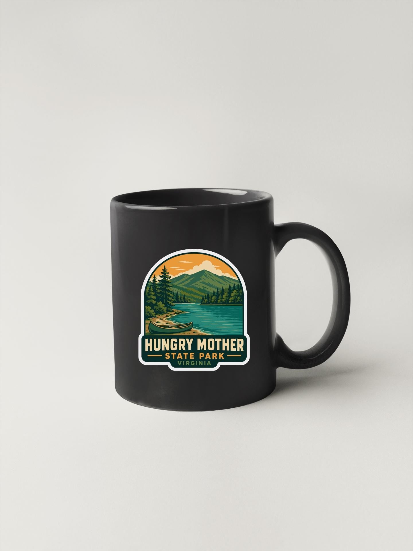 A black coffee mug with a graphic design depicting a scenic landscape, including mountains, trees, and a lake. The text on the mug reads "hungry mother state park".