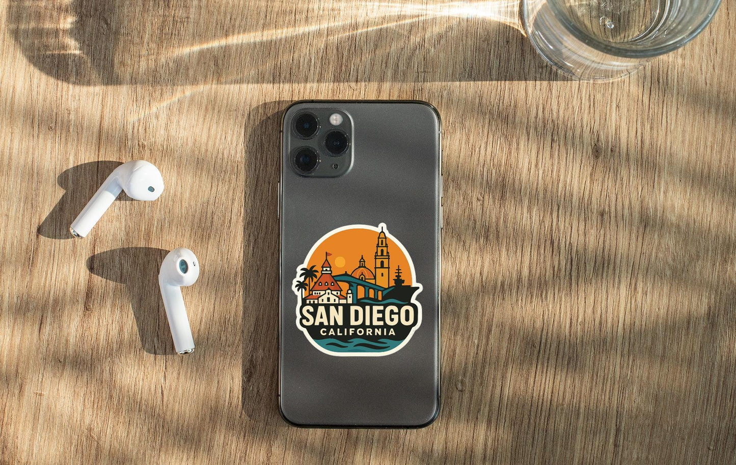 a smartphone with a sticker of San Diego, California, placed on a wooden surface alongside a pair of white earbuds and a glass of water.