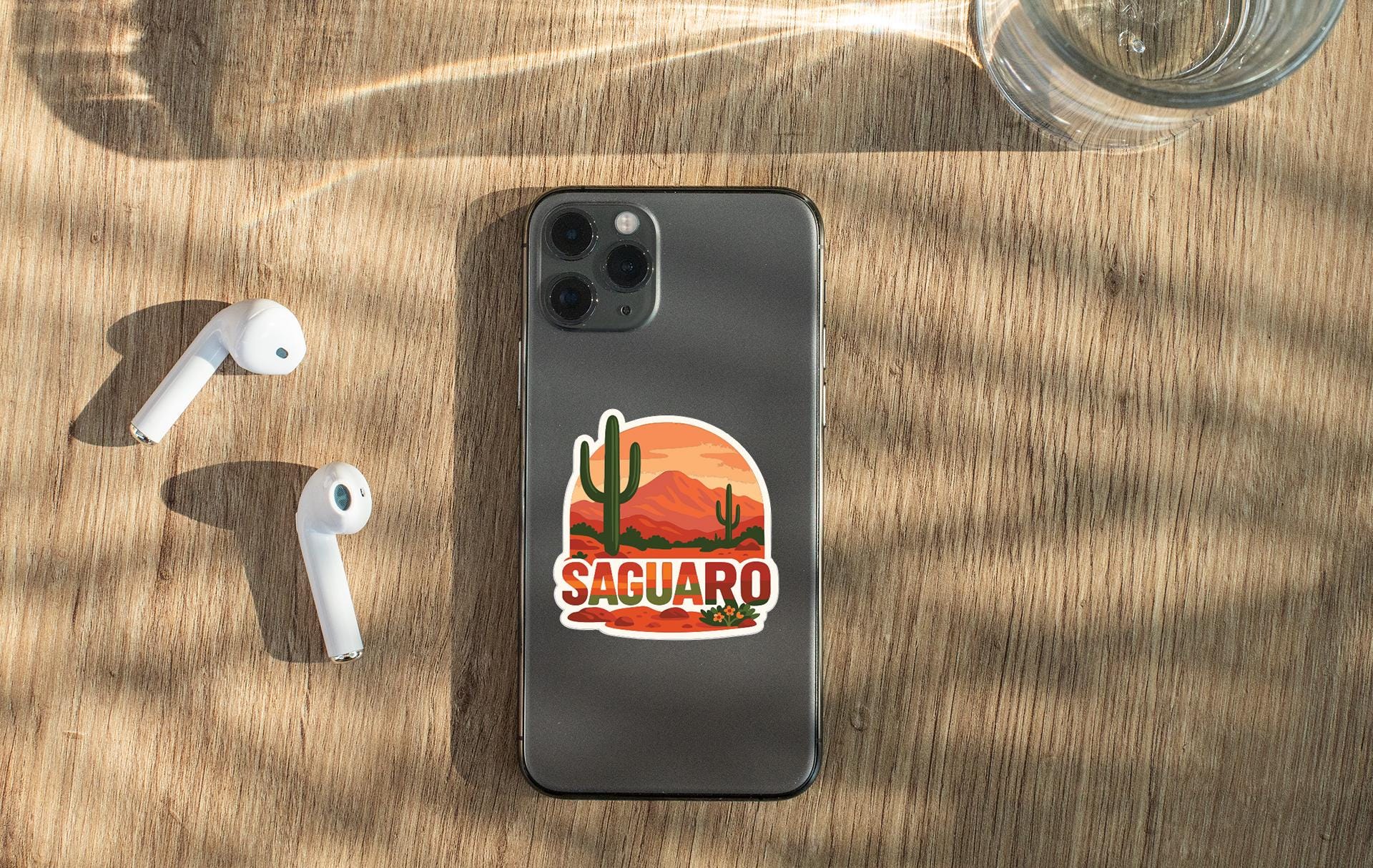 a smartphone with a sticker of a desert landscape, along with a pair of white earbuds and a glass of water on a wooden surface.