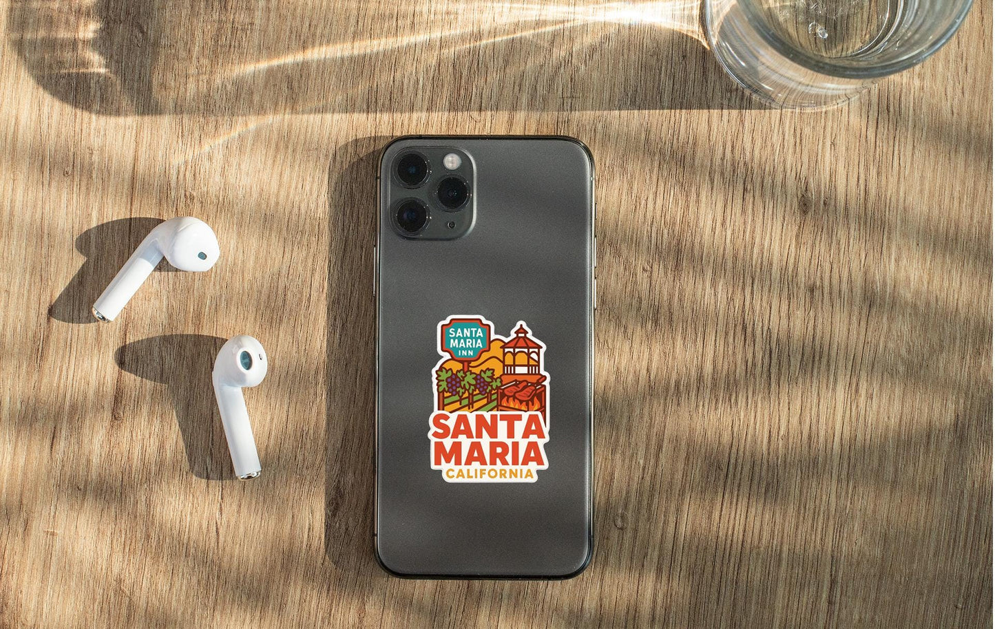 a smartphone with a sticker on the back, placed on a wooden surface alongside a pair of white earbuds and a glass of water.