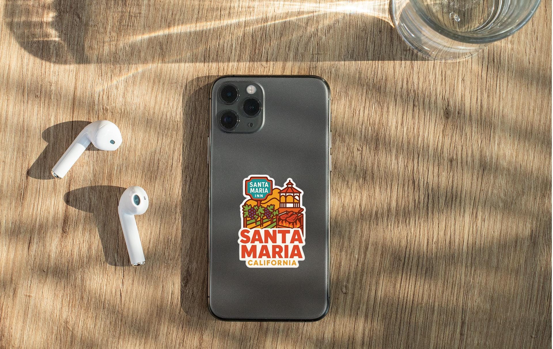 a smartphone with a sticker on the back, placed on a wooden surface alongside a pair of white earbuds and a glass of water.