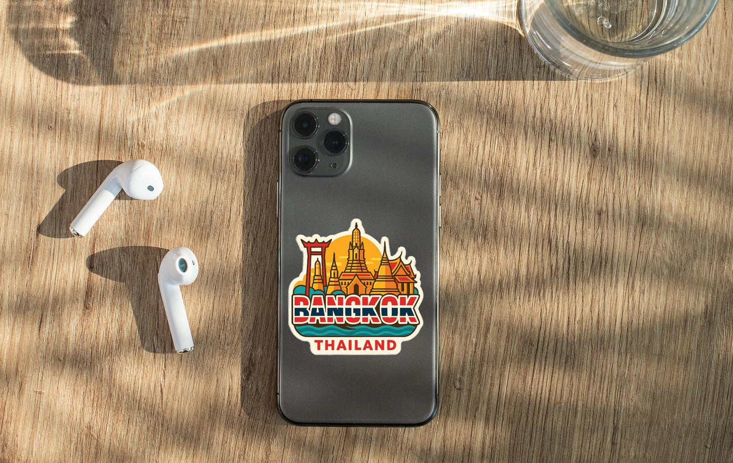 a smartphone with a sticker of Bangkok, Thailand, placed on a wooden surface alongside a pair of white earbuds and a glass of water.