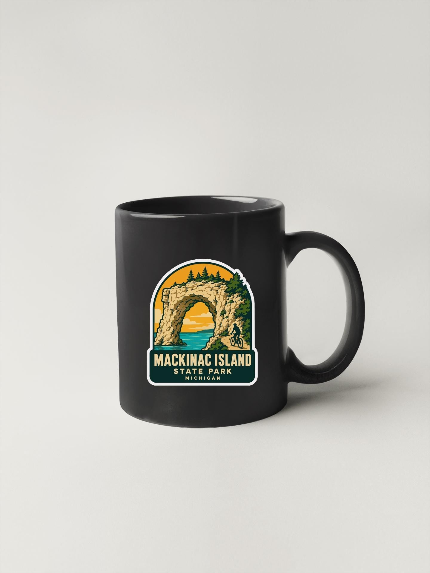 A black coffee mug with a graphic design depicting Mackinac Island, Michigan, featuring a large arch and a sunset over the water.