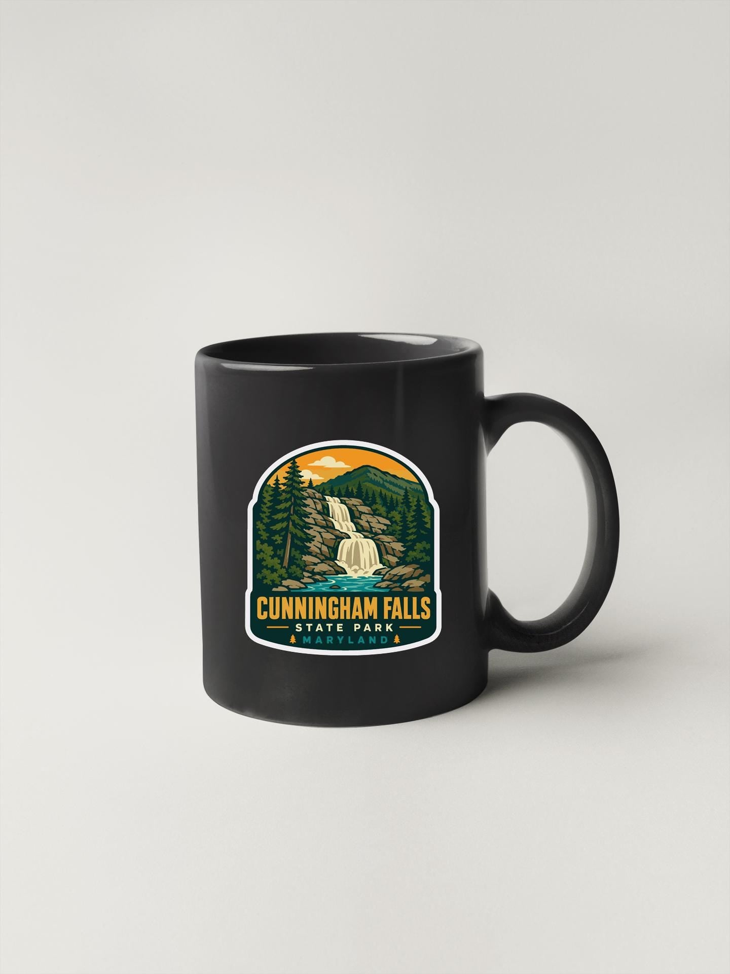 A black coffee mug with a graphic design depicting a waterfall and forest landscape, along with the text "CUNNINGHAM FALLS STATE PARK".