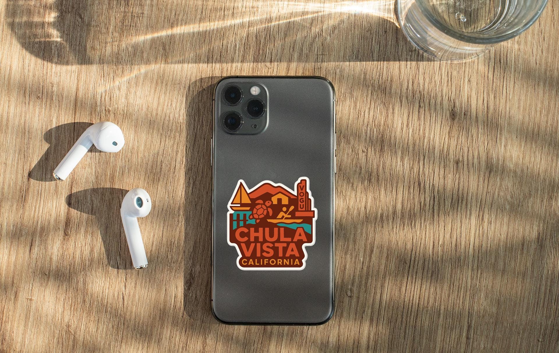 a smartphone with a sticker on the back, placed on a wooden surface alongside a pair of white earbuds and a glass of water.
