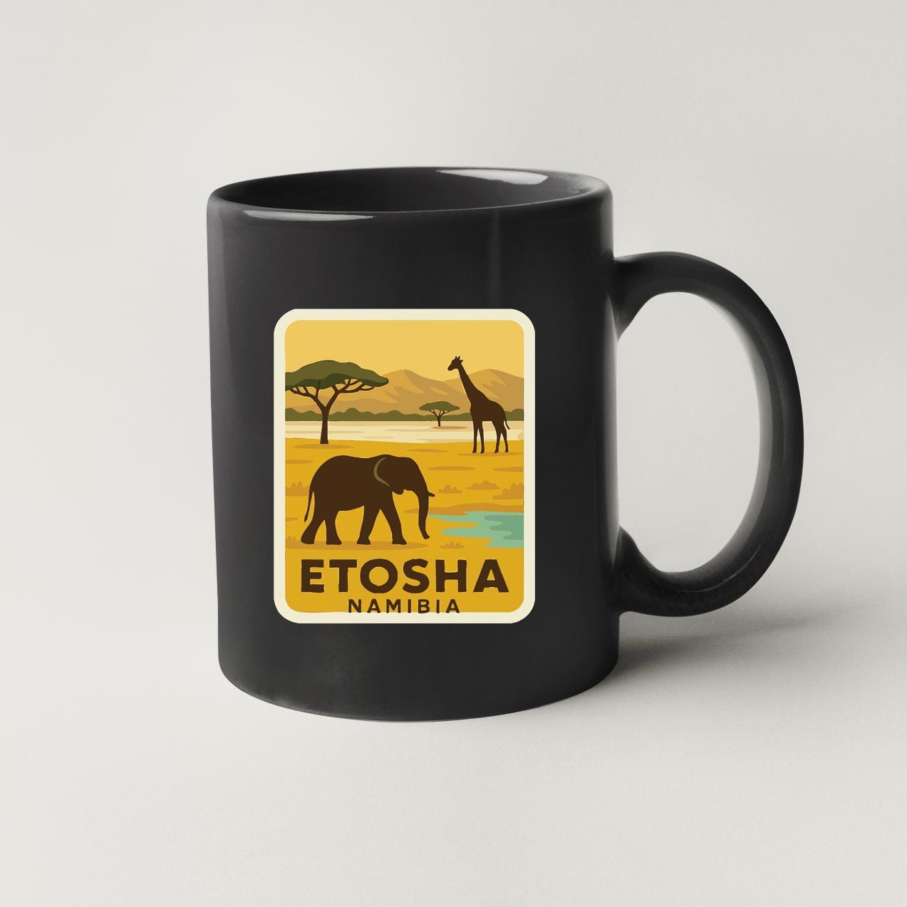 A black coffee mug with a yellow and green sticker depicting an elephant and a giraffe in a savanna landscape, along with the text "ETOSHA NAMIBIA".