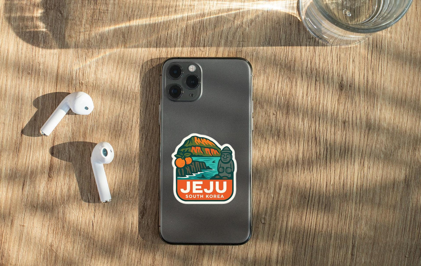 a smartphone with a sticker on the back, placed on a wooden surface alongside a pair of white earbuds and a glass of water.