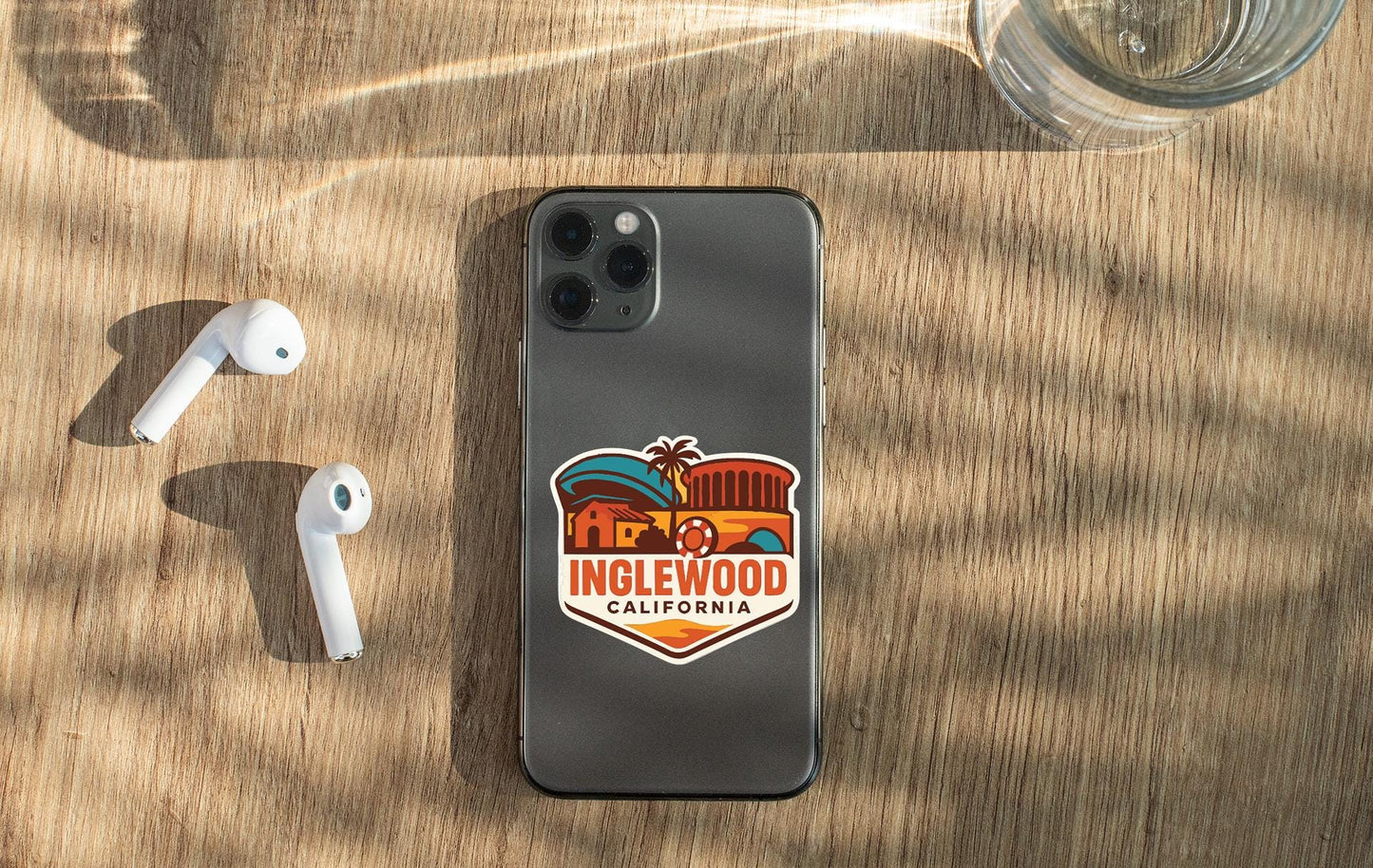a smartphone with a sticker on the back, placed on a wooden surface alongside a pair of white earbuds and a glass of water.