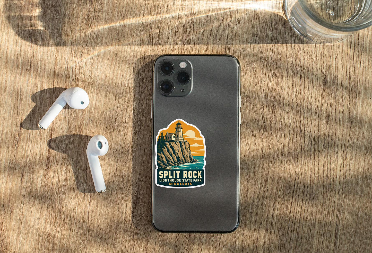 a smartphone with a sticker on it, placed on a wooden surface alongside a pair of white earbuds and a glass of water.
