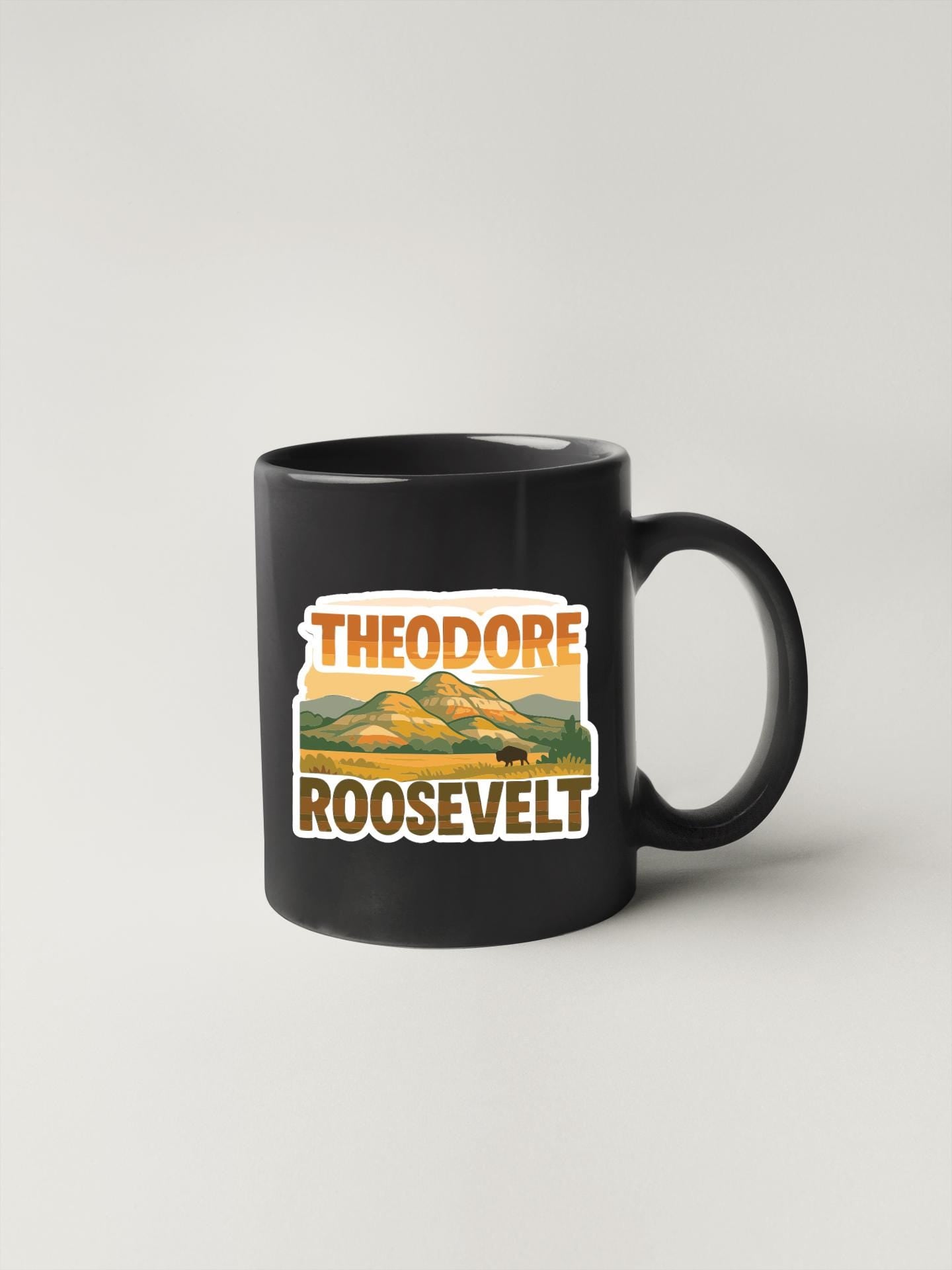A black coffee mug with a sticker featuring the text "THEODORE ROOSEVELT" and an illustration of a mountain landscape.