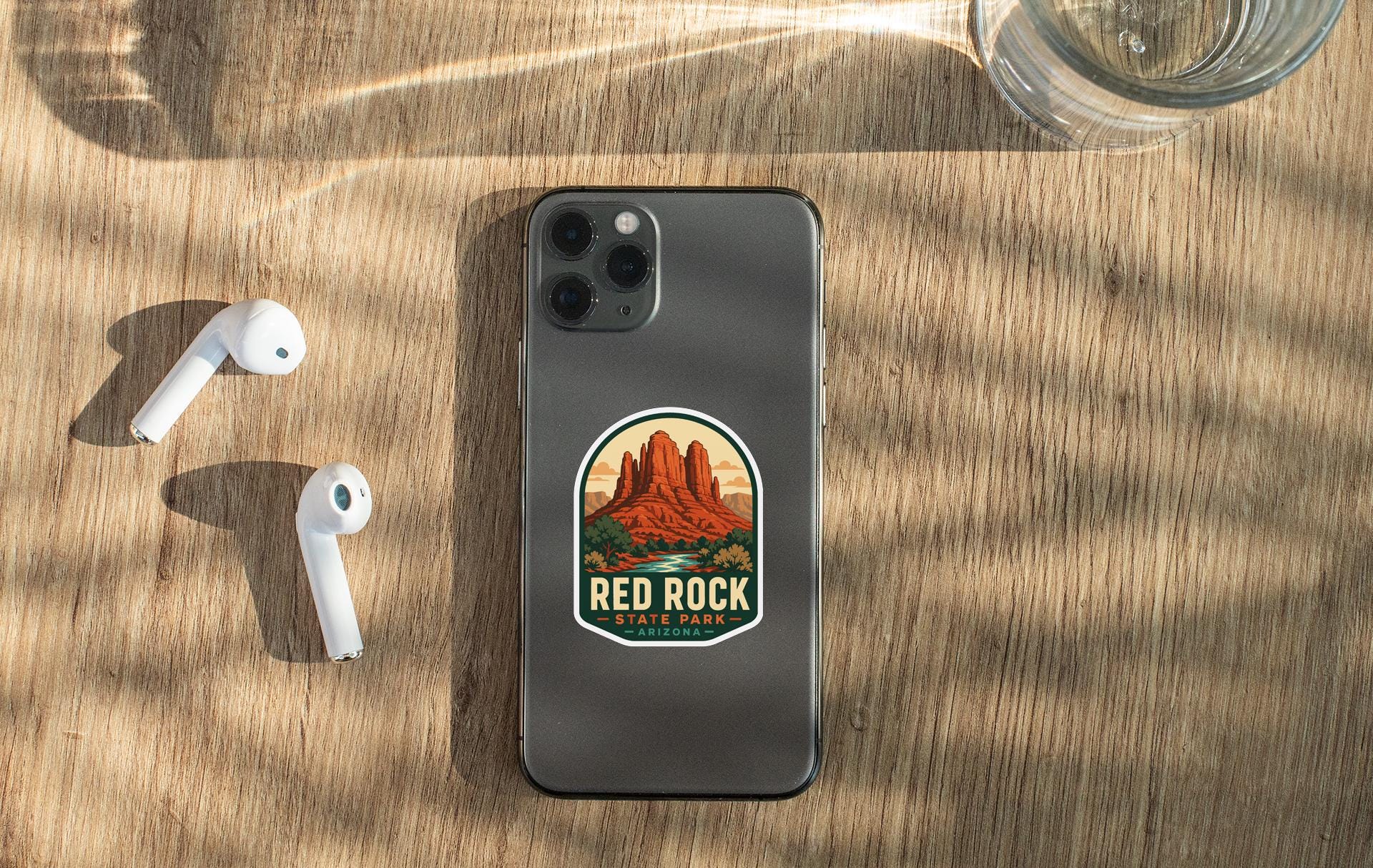 a smartphone with a sticker of Red Rock State Park on its back, along with a pair of white earbuds and a glass of water on a wooden surface.