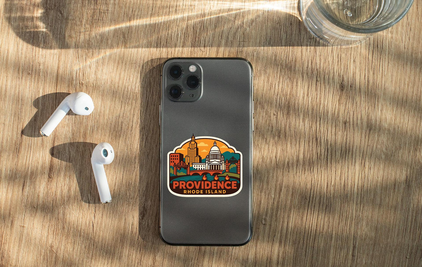 a smartphone with a sticker of Providence Rhode Island on its back, along with a pair of white earbuds and a glass of water on a wooden surface.