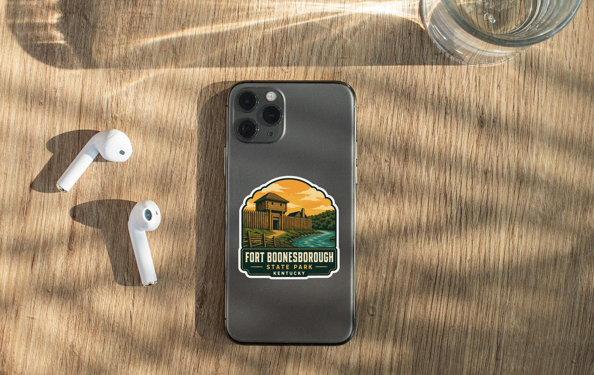 a smartphone with a sticker on the back, placed on a wooden surface alongside a pair of white earbuds and a glass of water.