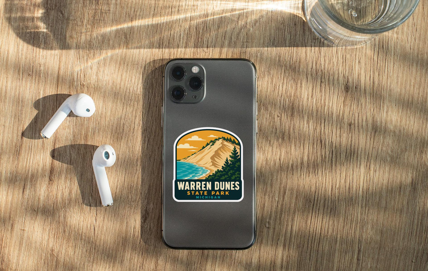 a smartphone with a sticker on it, placed on a wooden surface alongside a pair of white earbuds and a glass of water.