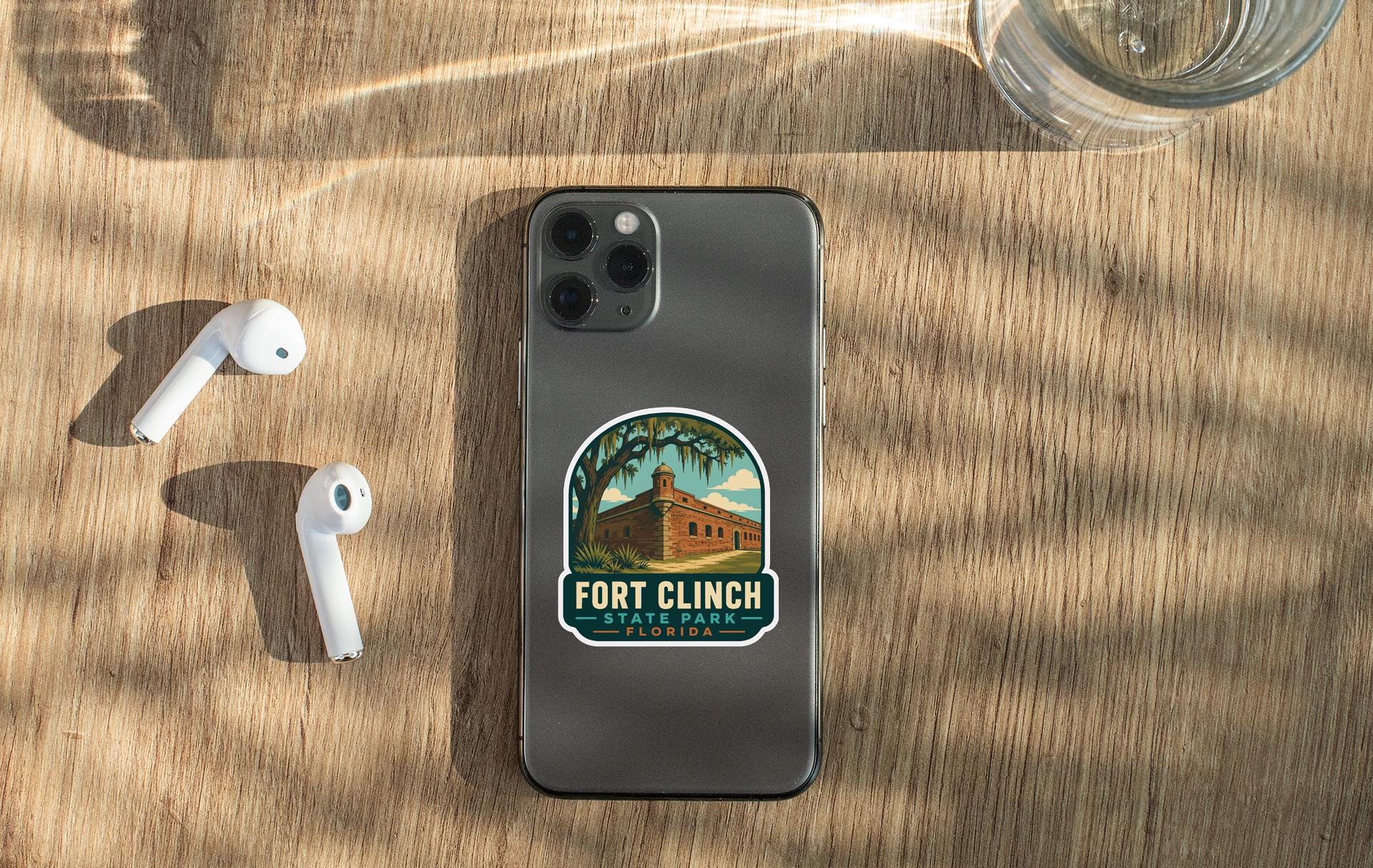 a smartphone with a sticker on the back, placed on a wooden surface alongside a pair of white earbuds and a glass of water.