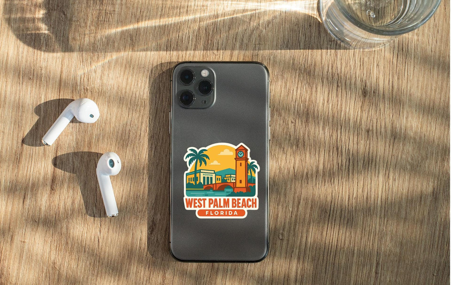 a smartphone with a sticker of West Palm Beach, Florida, placed on a wooden surface alongside a pair of white earbuds and a glass of water.