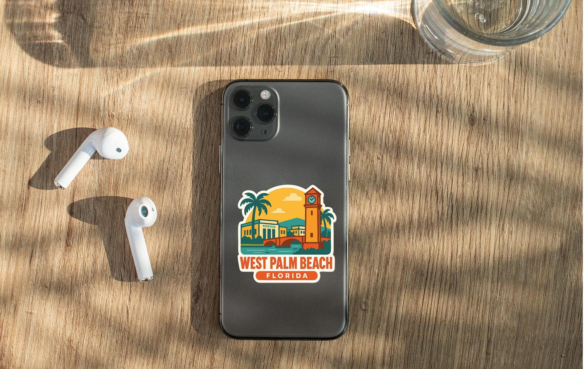 a smartphone with a sticker of West Palm Beach, Florida, placed on a wooden surface alongside a pair of white earbuds and a glass of water.