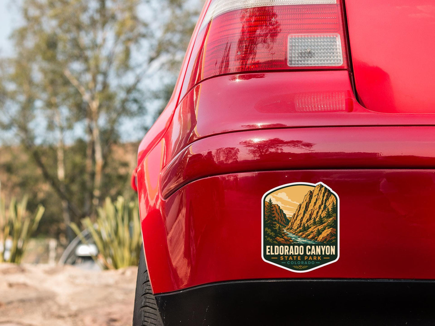 the rear end of a red car with a sticker on the back, featuring a scenic mountain landscape.