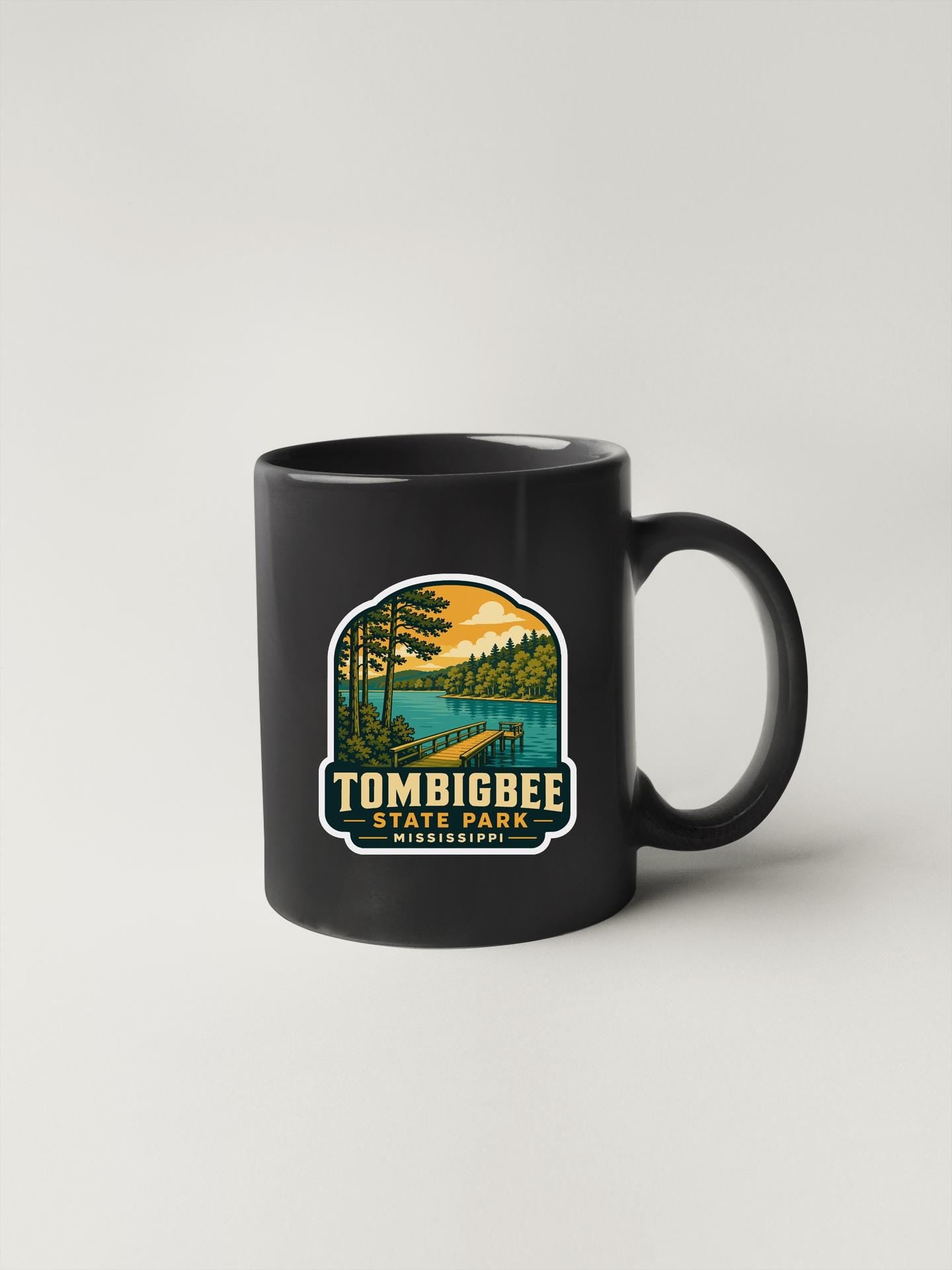 A black coffee mug with a graphic design depicting a scenic landscape, including a lake, trees, and a bridge.