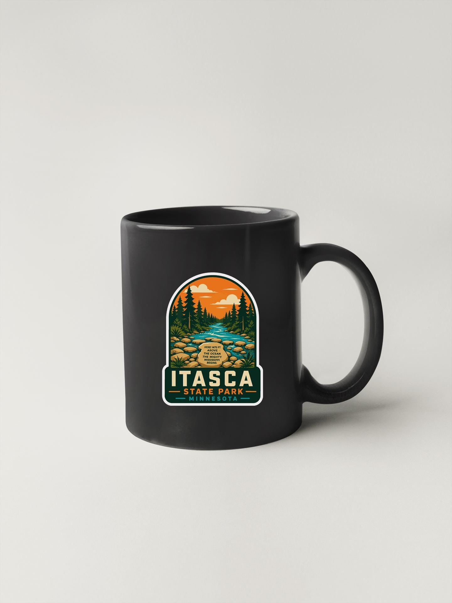 A black coffee mug with a graphic design depicting a scenic landscape with pine trees, a river, and a sunset.
