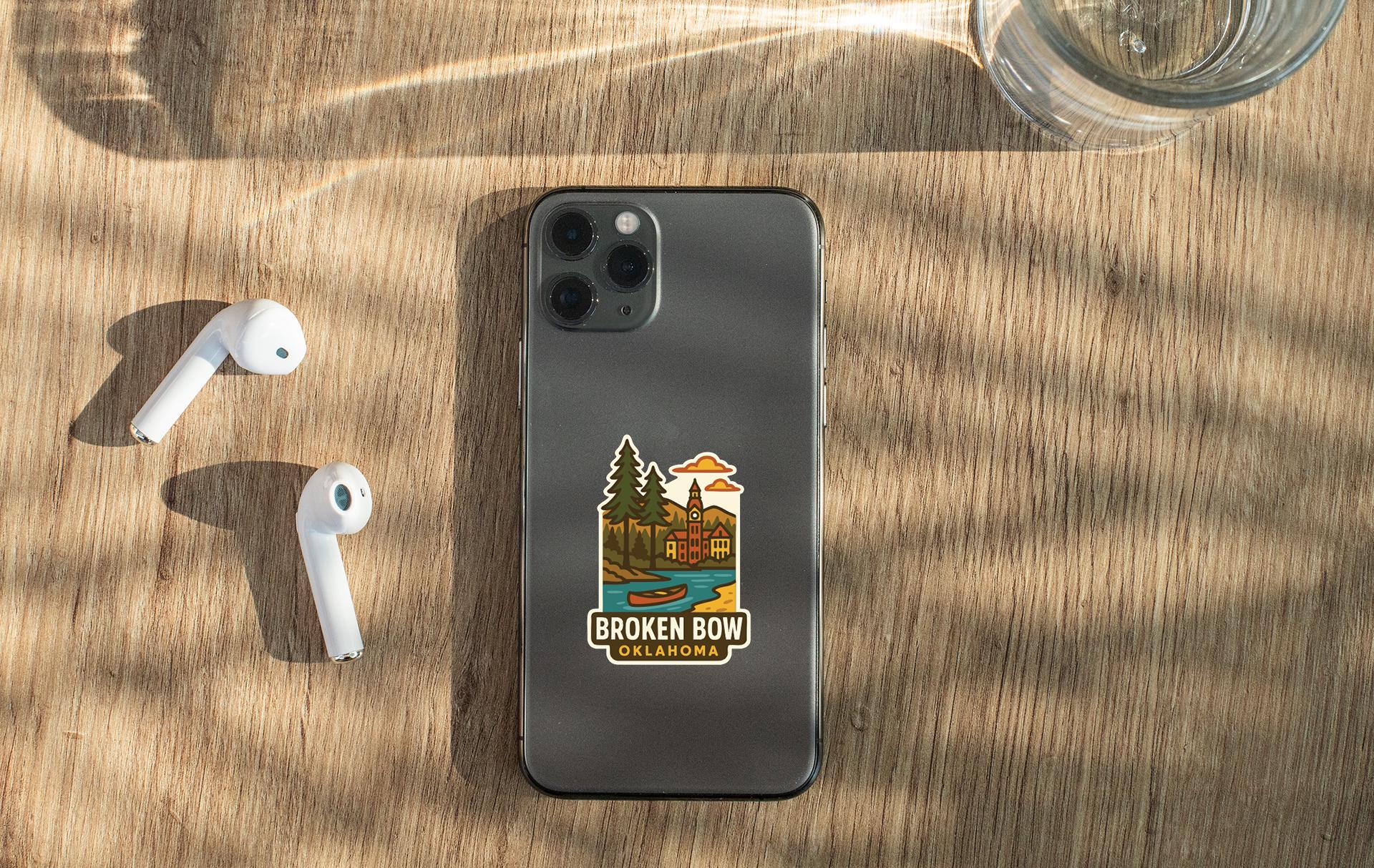 a smartphone with a sticker on the back, placed on a wooden surface alongside a pair of white earbuds and a glass of water.