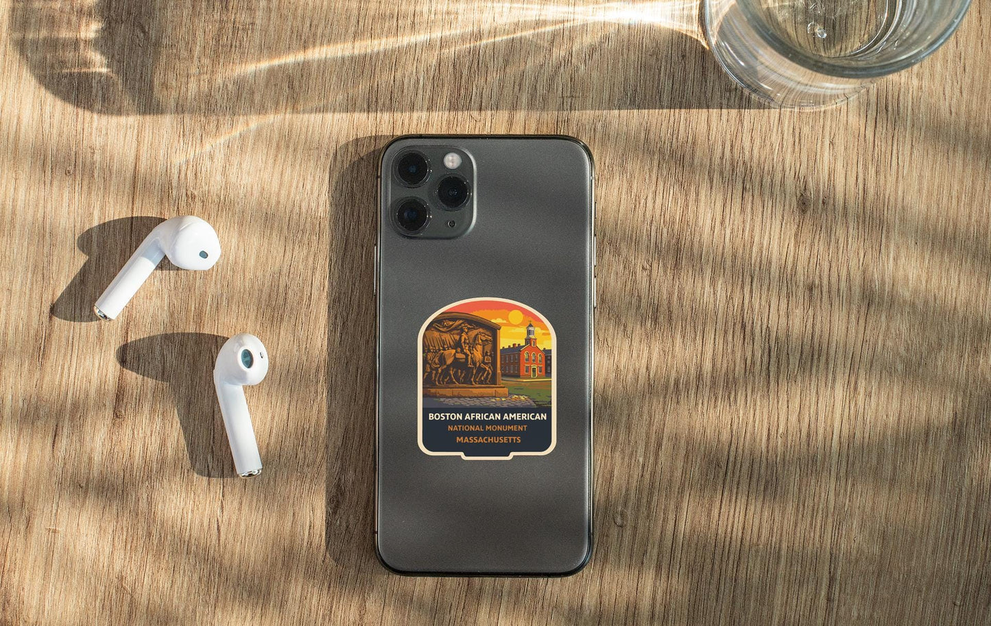 a smartphone with a sticker on the back, placed on a wooden surface alongside a pair of white earbuds and a glass of water.