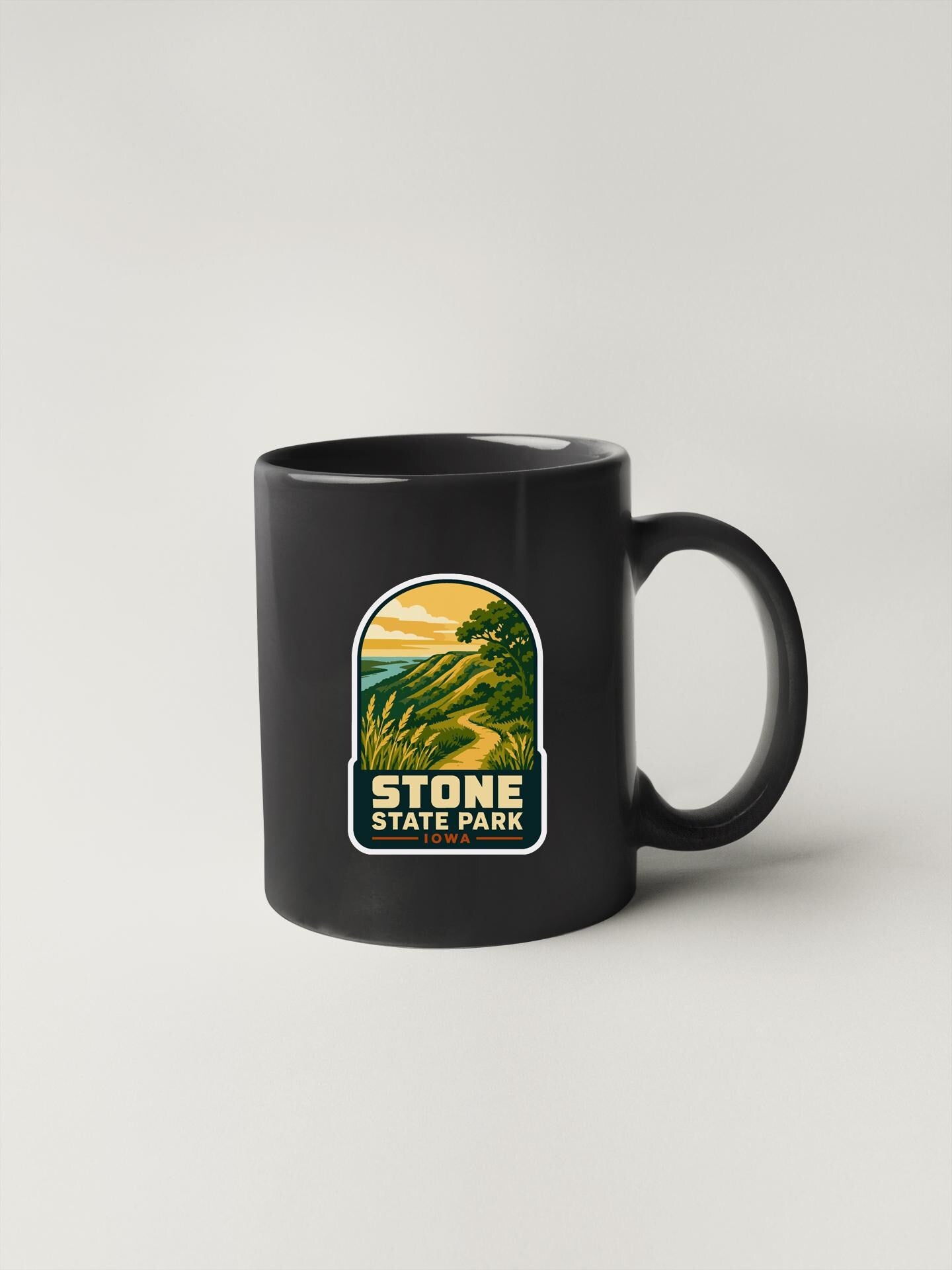 A black coffee mug with a graphic design depicting a scenic landscape, featuring a mountain range and a winding path.