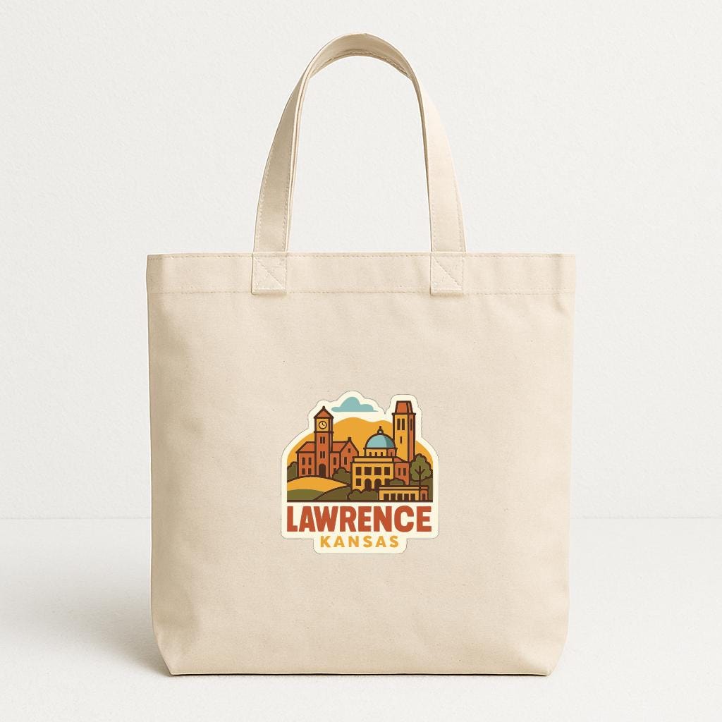 A beige tote bag with a printed design of a cityscape and the text "LAWRENCE KANSAS" printed on it.