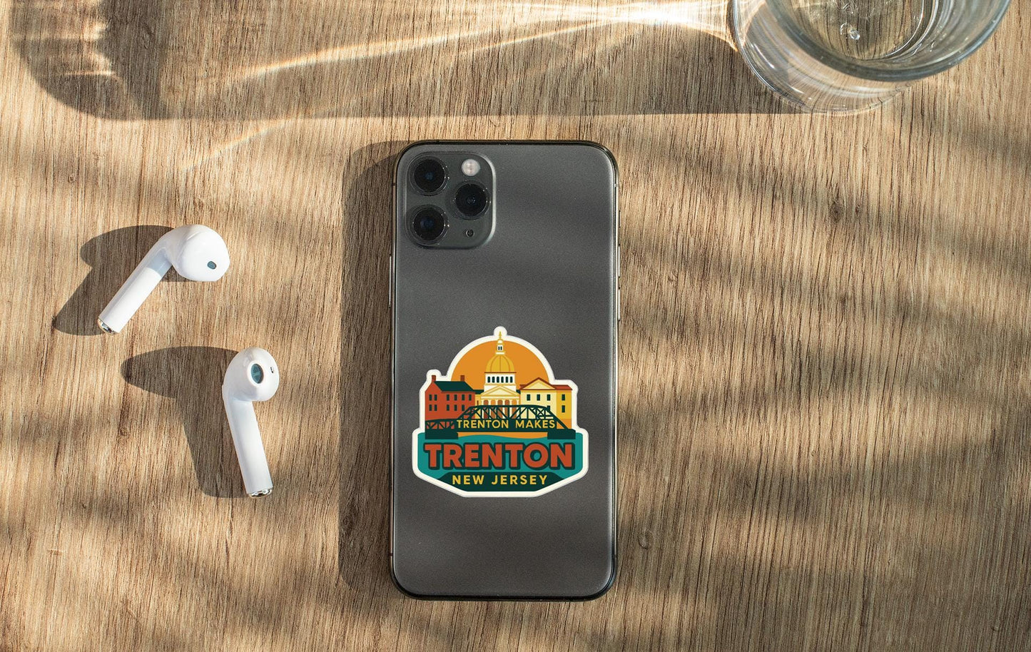 a smartphone with a sticker on it, placed on a wooden surface alongside a pair of white earbuds and a glass of water.