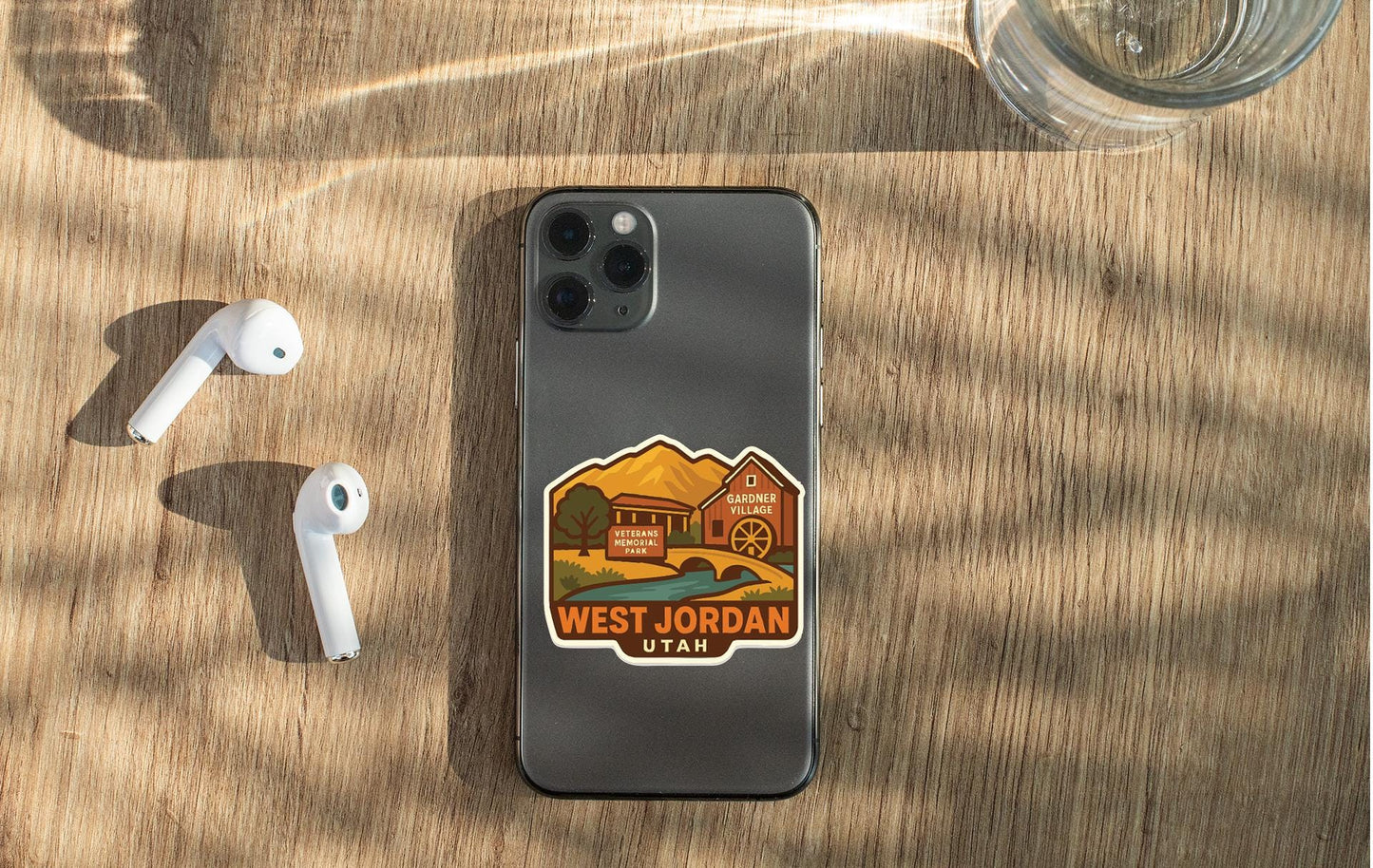 a smartphone with a sticker on the back, placed on a wooden surface alongside a pair of white earbuds and a glass of water.