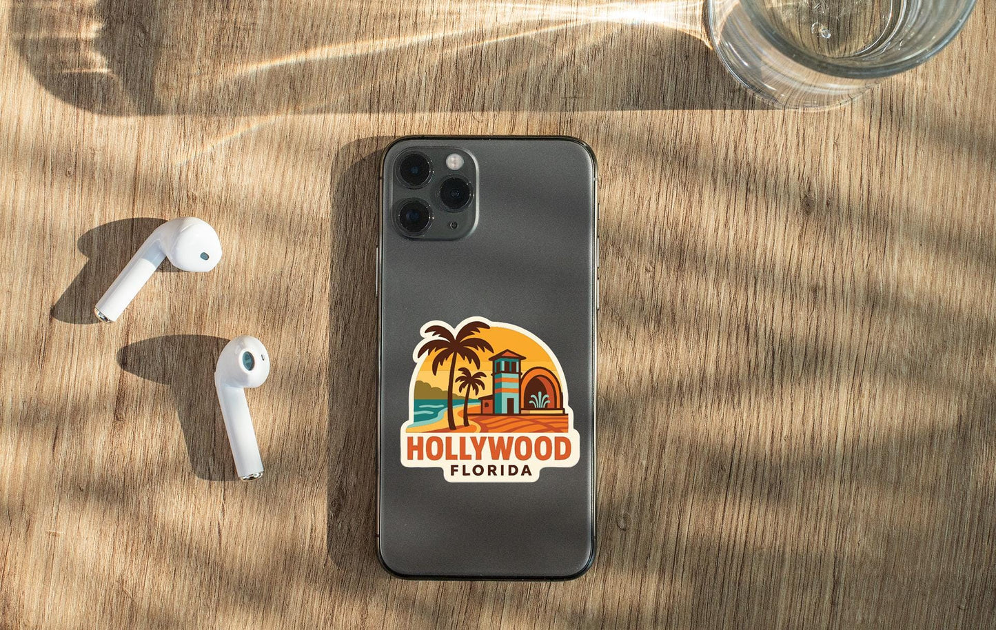 a smartphone with a sticker of Hollywood, Florida on its back, along with a pair of white earbuds and a glass of water on a wooden surface.