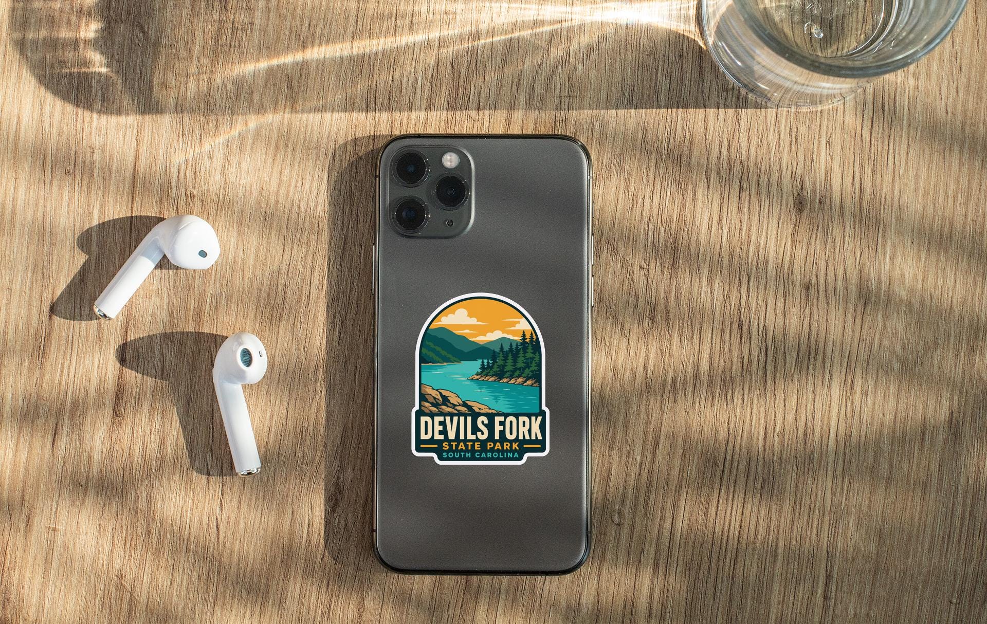 a smartphone with a sticker on it, placed on a wooden surface alongside a pair of white earbuds and a glass of water.