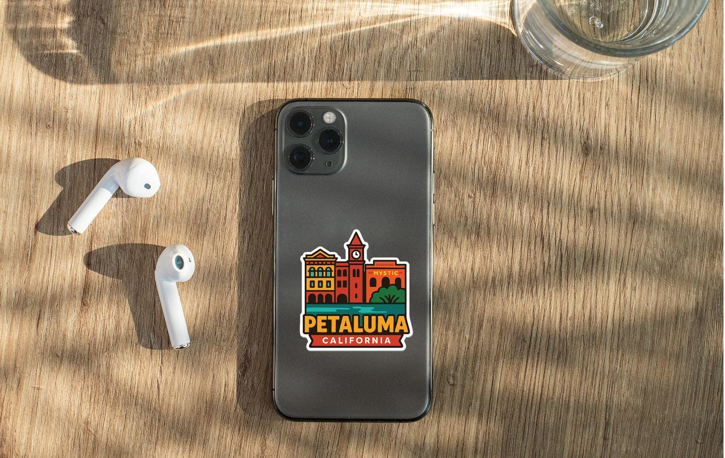 a smartphone with a sticker of a city on it, placed on a wooden surface alongside a pair of white earbuds and a glass of water.