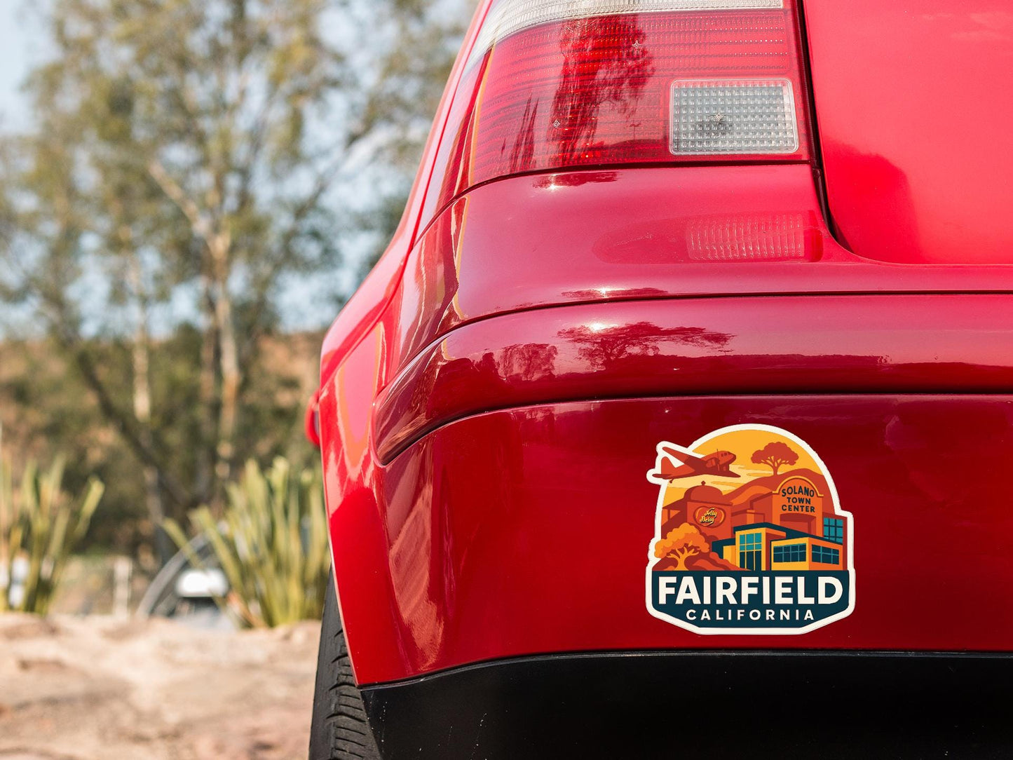 the rear end of a red car with a sticker on it, featuring a scenic illustration of a town or city.