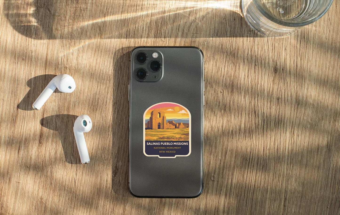 a smartphone with a sticker on the back, placed on a wooden surface alongside a pair of white earbuds and a glass of water.