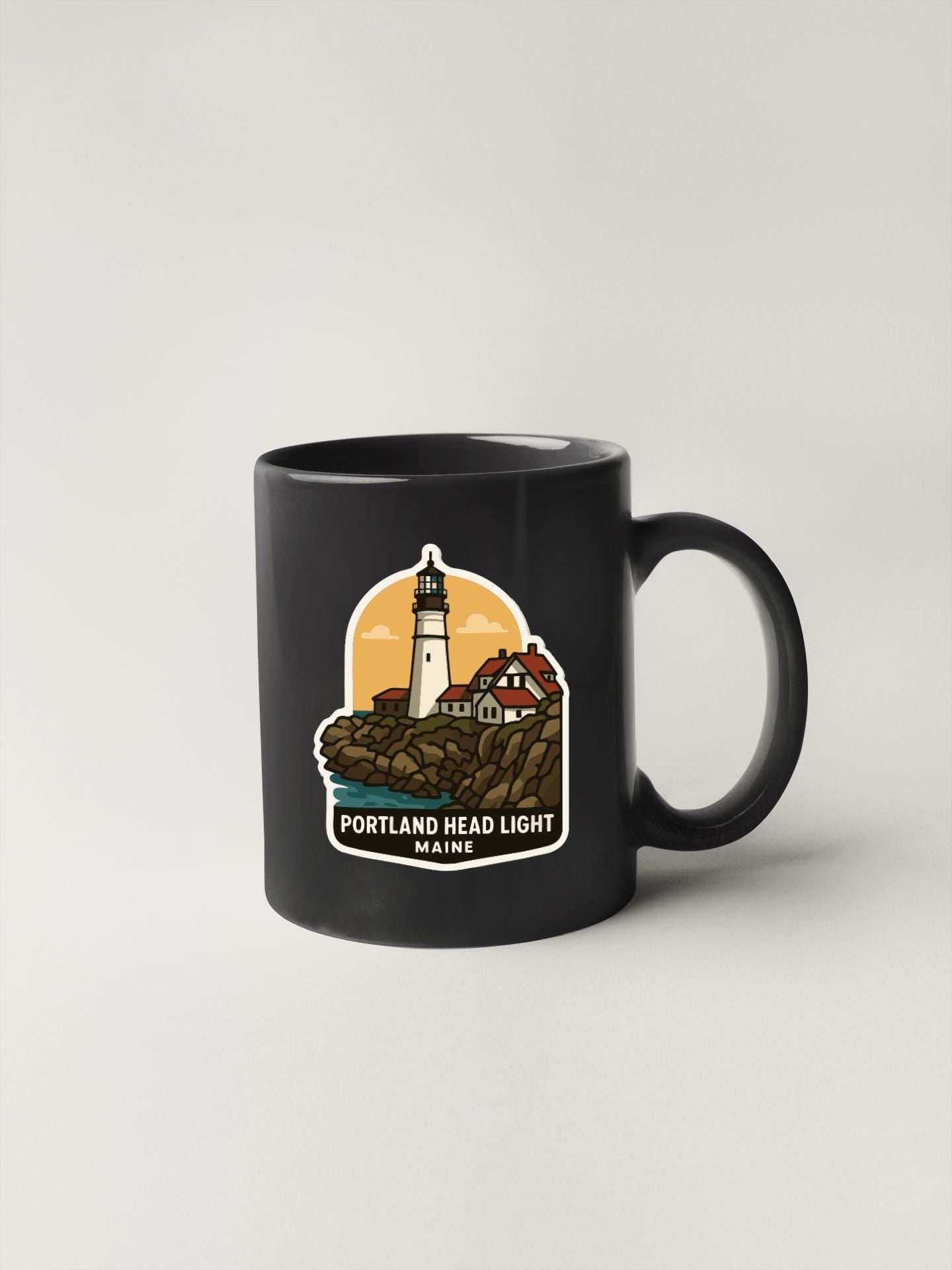 A black coffee mug with a graphic design depicting a lighthouse and a house, along with the text "PORTLAND HEAD LIGHT MAINE".