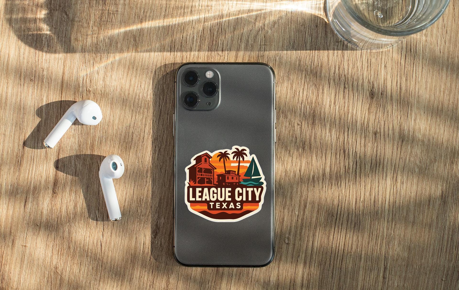 a smartphone with a sticker on the back, placed on a wooden surface alongside a pair of white earbuds and a glass of water.